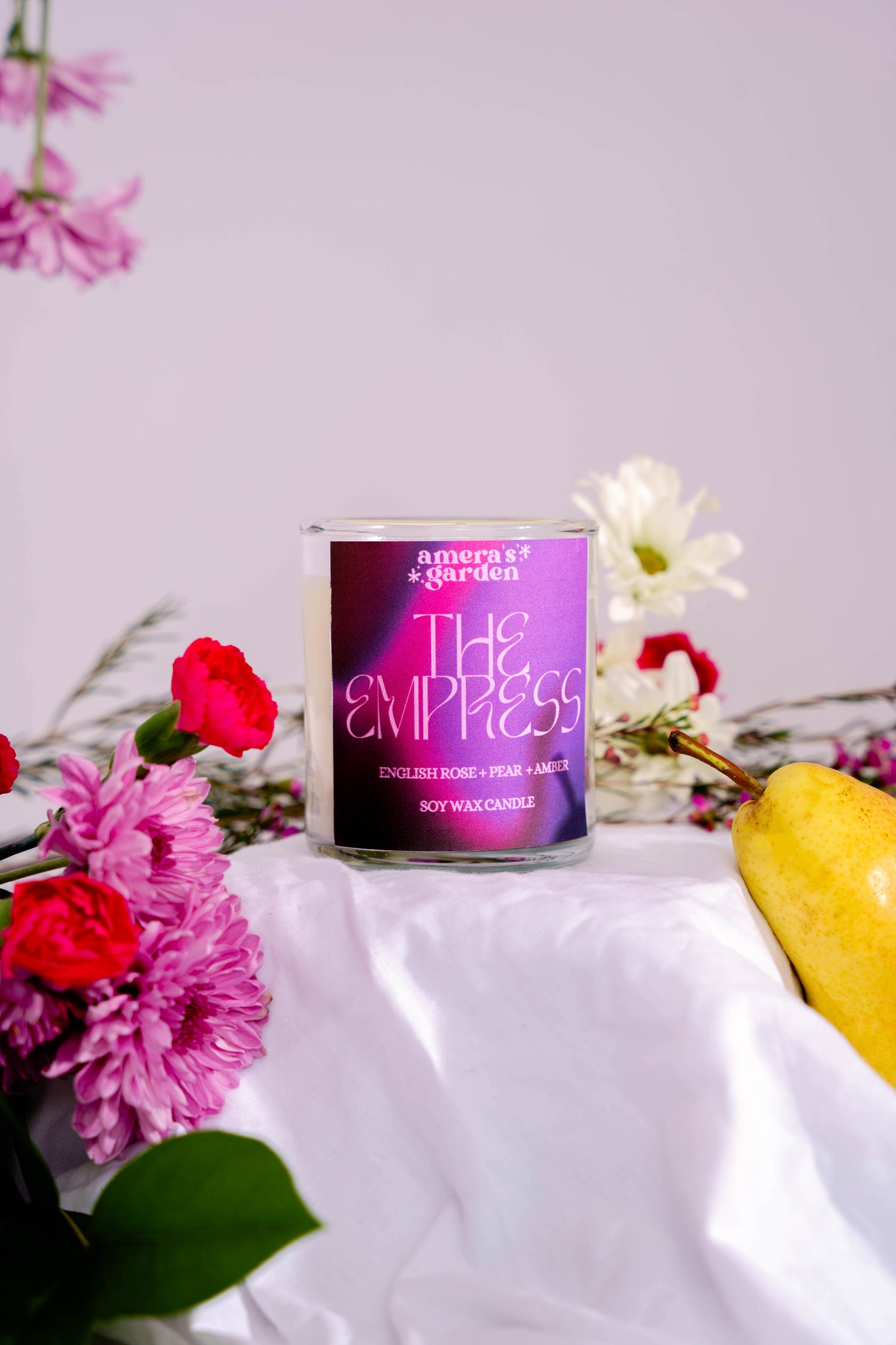 A purple candle with the label 'The Empress' by Amera's Garden, surrounded by flowers and a banana, on a light background.