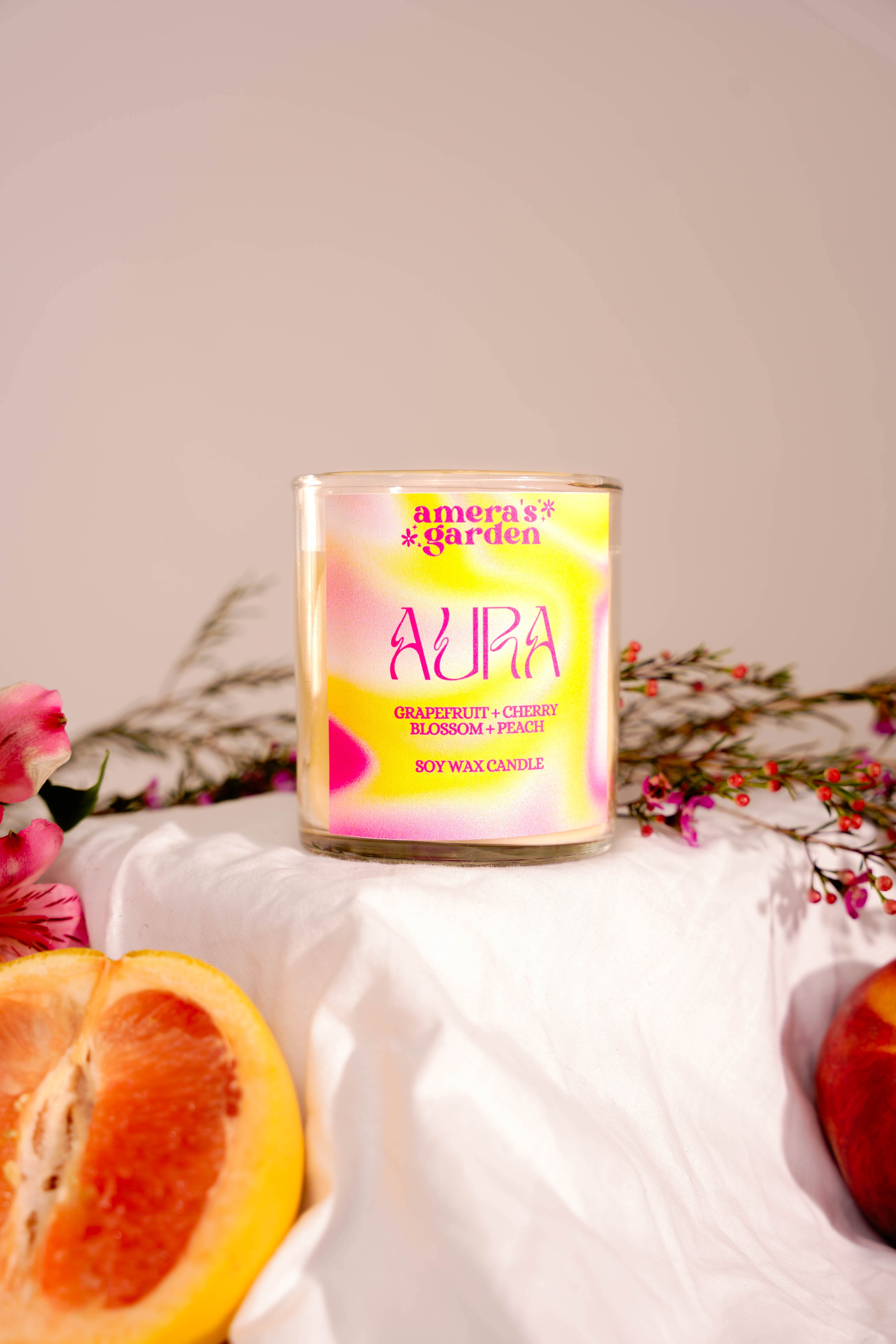 A peach-colored candle with a clear glass container, labeled 'AURA' and described as a 'Royalty Blend' with notes of peach, mandarin, and teakwood, presented on a white surface with floral decor in the background.