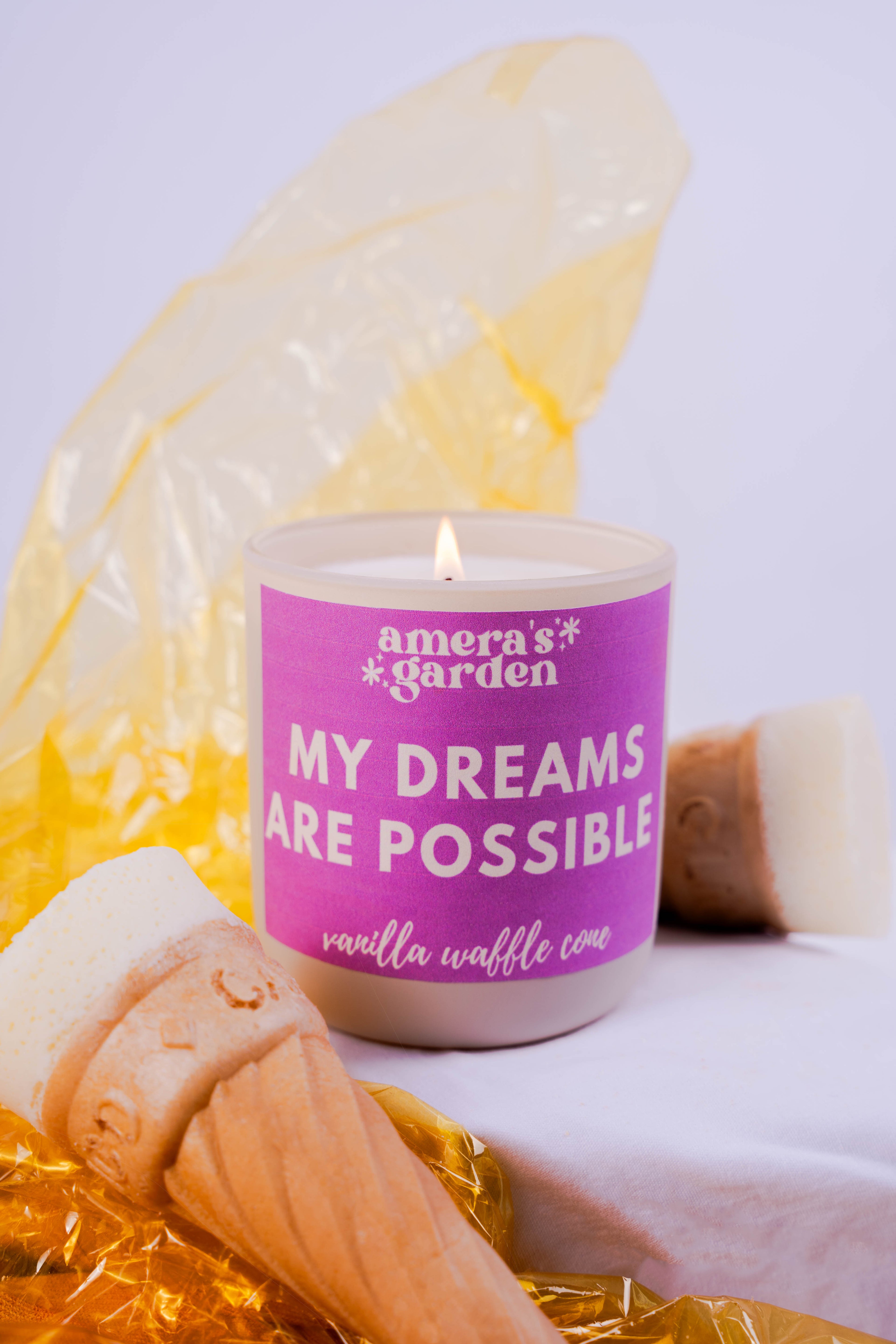A purple candle with white text that reads 'Amera's Garden - My Dreams Are Possible' alongside a yellow waffle cone.