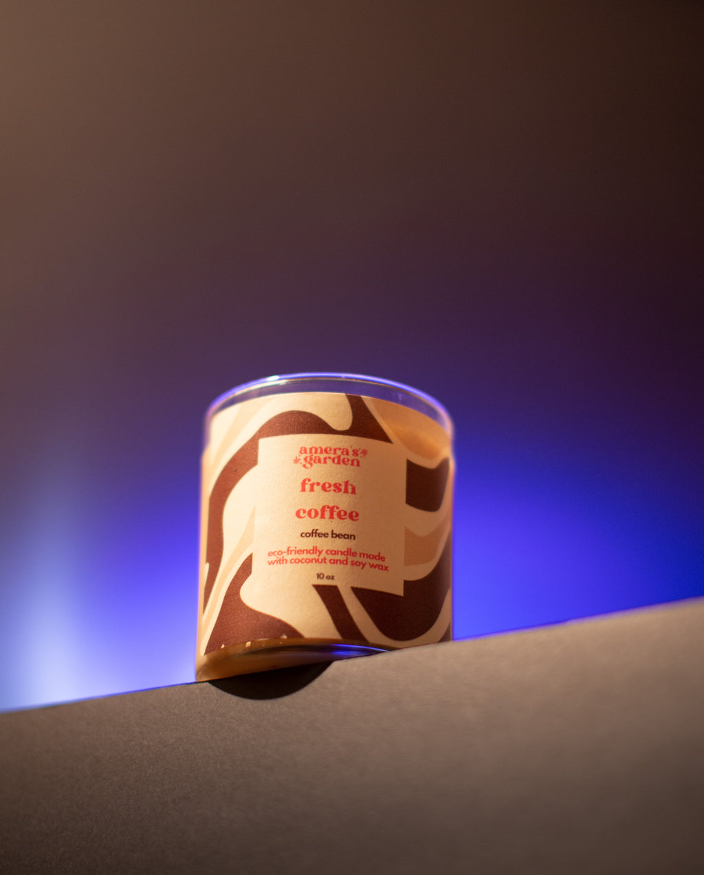 A jar of Fresh Coffee candle with a brown label, placed on a table with a cup of coffee and a teapot in the background, suggesting a theme of coffee lovers.