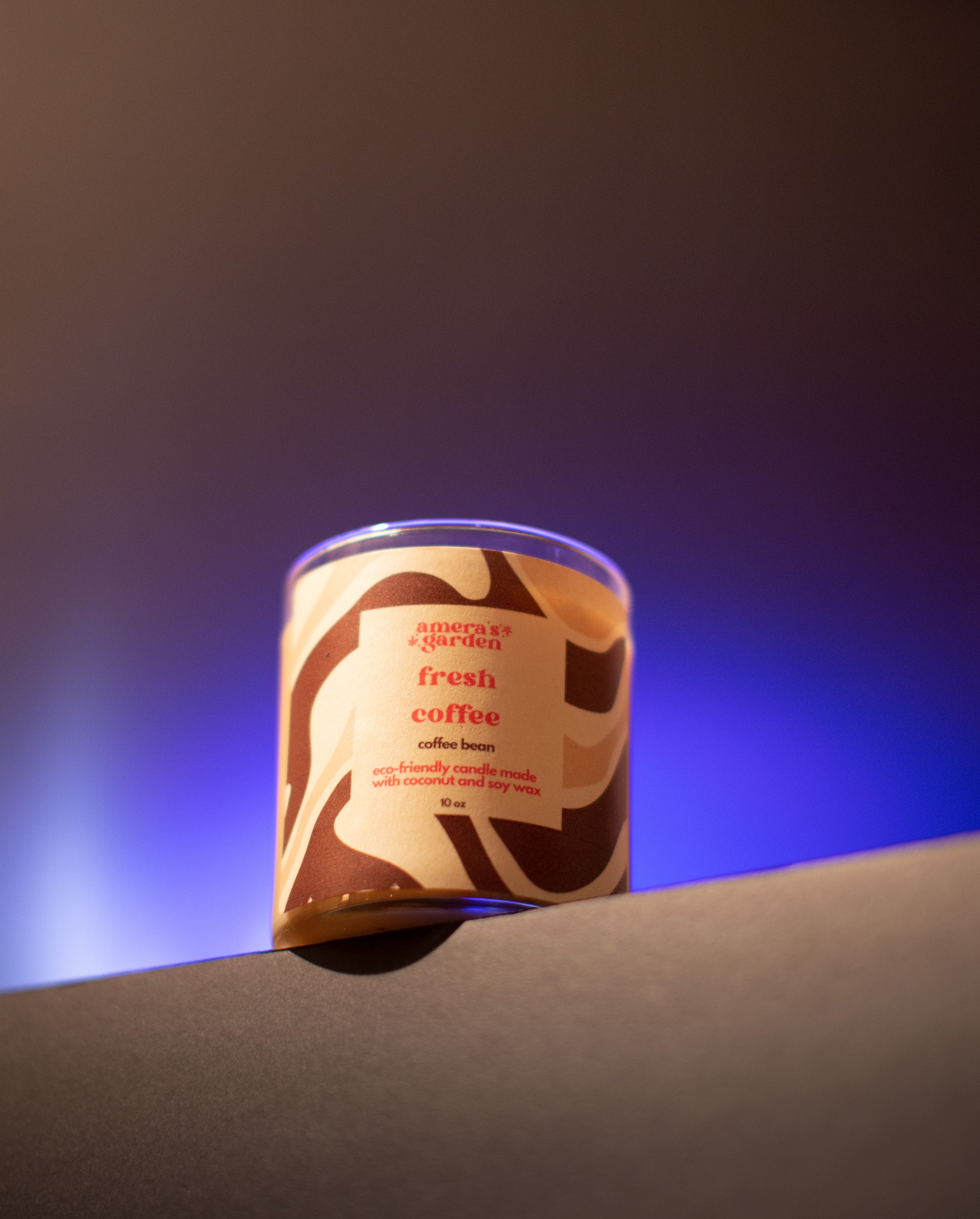 A jar of Fresh Coffee candle with a brown label, placed on a table with a cup of coffee and a teapot in the background, suggesting a theme of coffee lovers.