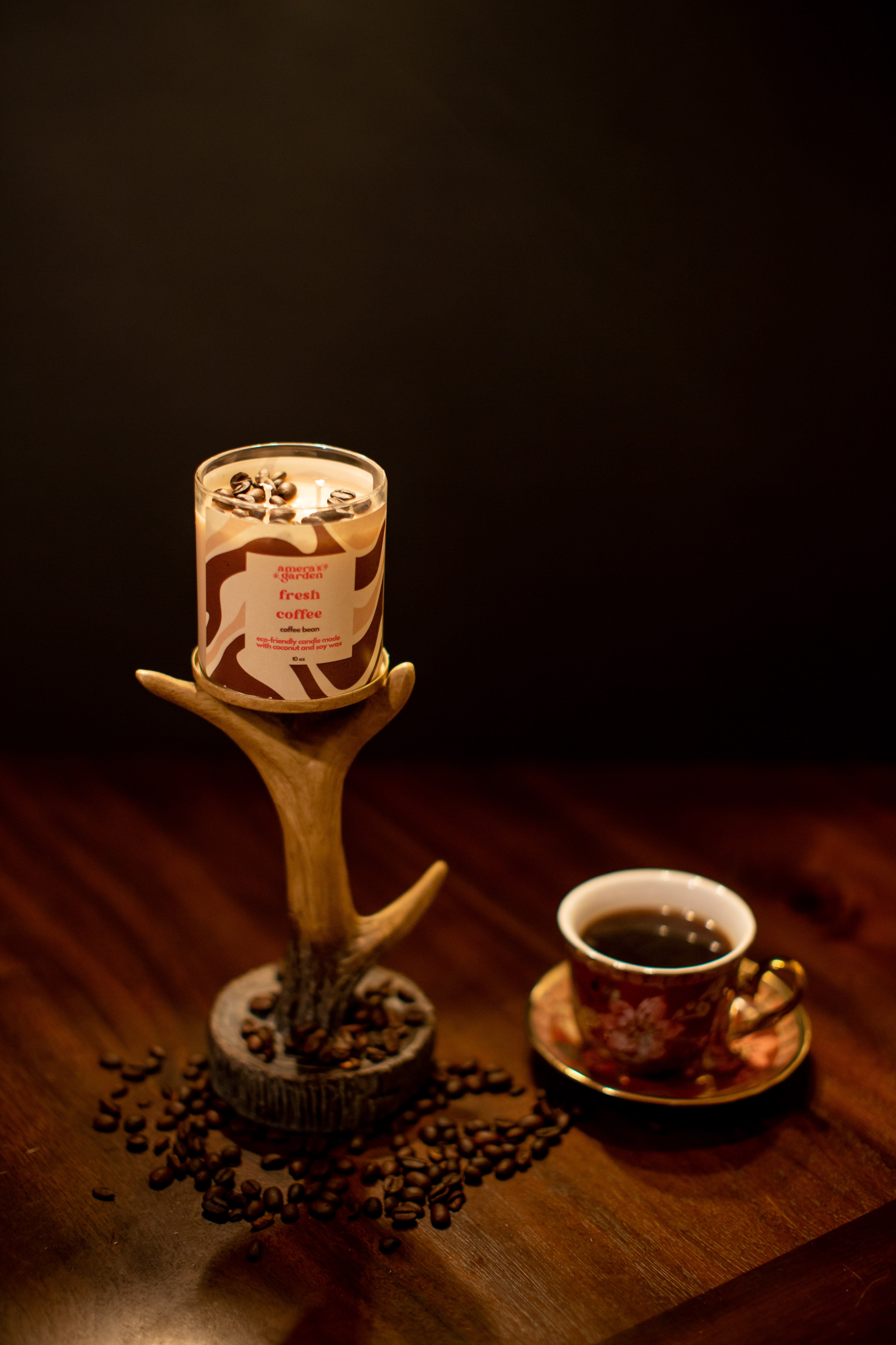 A jar of Fresh Coffee candle with a brown label, placed on a table with a cup of coffee and a teapot in the background, suggesting a theme of coffee lovers.